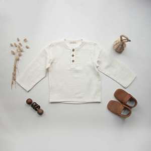 A neutral-toned boho baby boy shirt made from organic muslin fabric, featuring long sleeves and natural wooden buttons, displayed in a minimal nursery setting – perfect for summer wear or vacation outfits.