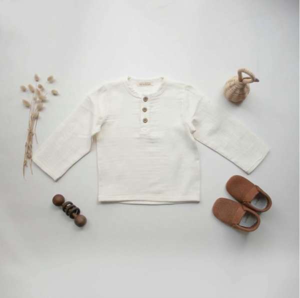 A neutral-toned boho baby boy shirt made from organic muslin fabric, featuring long sleeves and natural wooden buttons, displayed in a minimal nursery setting – perfect for summer wear or vacation outfits.