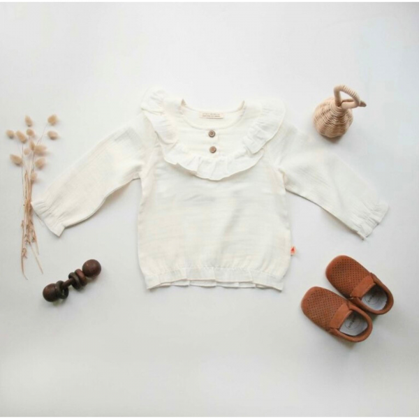 Weißes Baby-Oberteil aus Bio-Musselin mit langen Ärmeln, Rüschenkragen und Holzknöpfen, flach gestylt mit braunen Babyschuhen und getrockneten Blumen.
